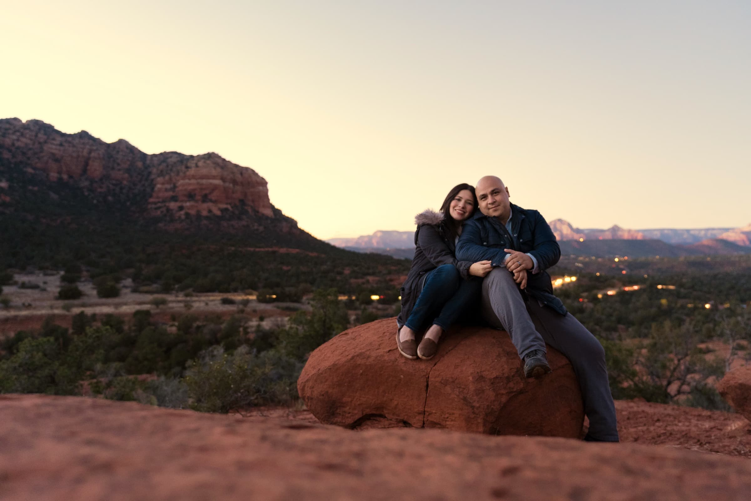 Mario and Brenda Silva sitting together on a red sandstone boulder at dusk, red rock formations behind them and the lights of a town coming on in the valley below