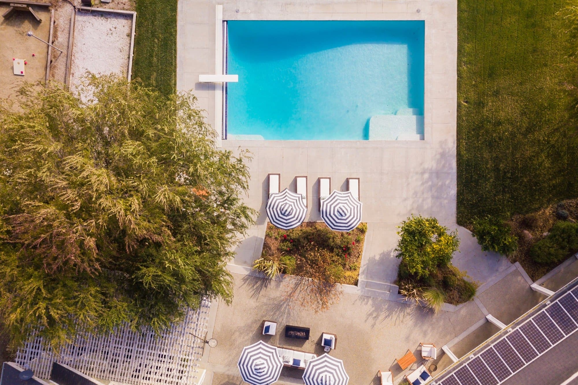 A property photographed from directly overhead: a rectangular pool, a run of loungers, three striped umbrellas, a planted bed and a lawn, all readable at once as a plan