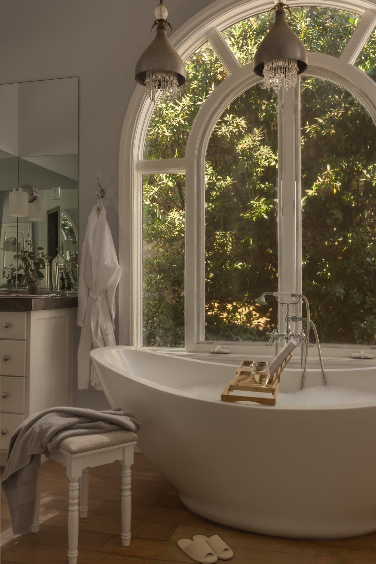 A freestanding tub beneath a tall arched window filled edge to edge with dense foliage.