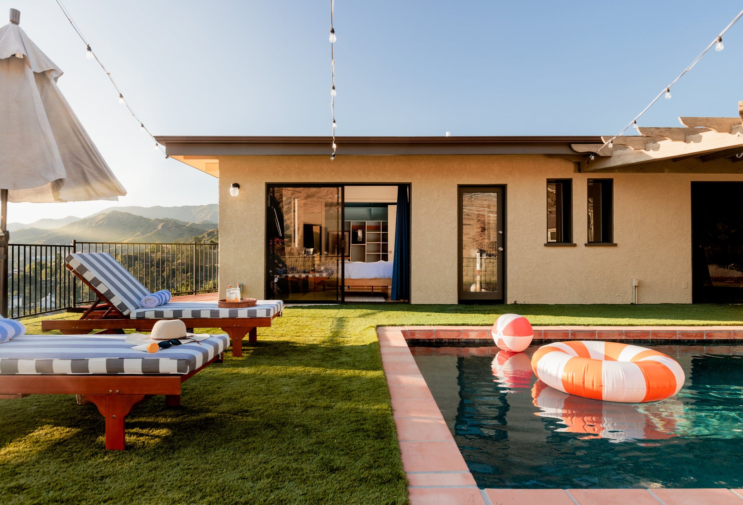 Low evening sun across a lawn and pool, with the house open to a bedroom, two striped loungers to one side, and mountains beyond the railing.