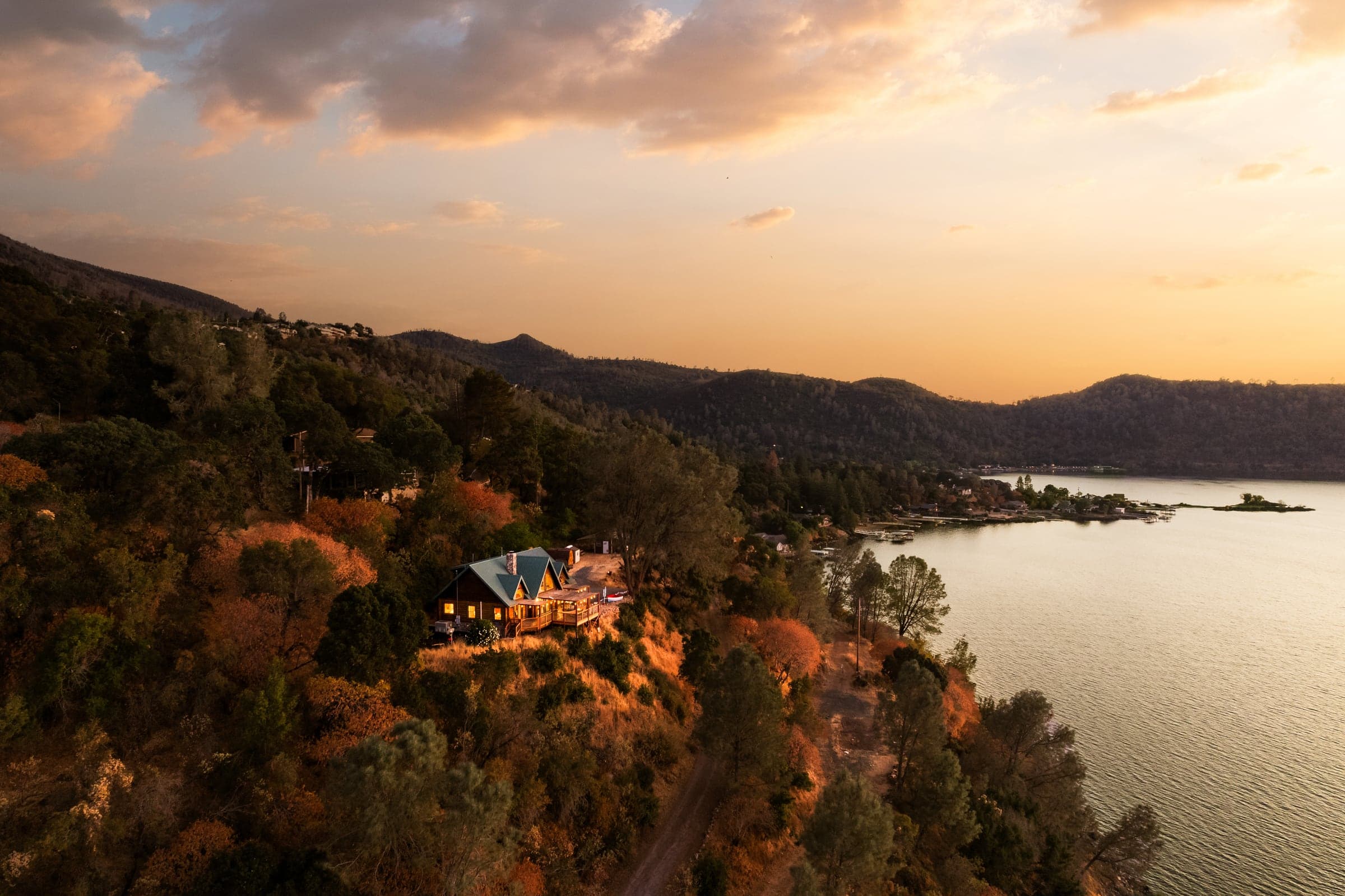 Aerial sunset view of a lit house on a wooded slope above Clear Lake, with shoreline homes, water, and ridgelines visible around it.