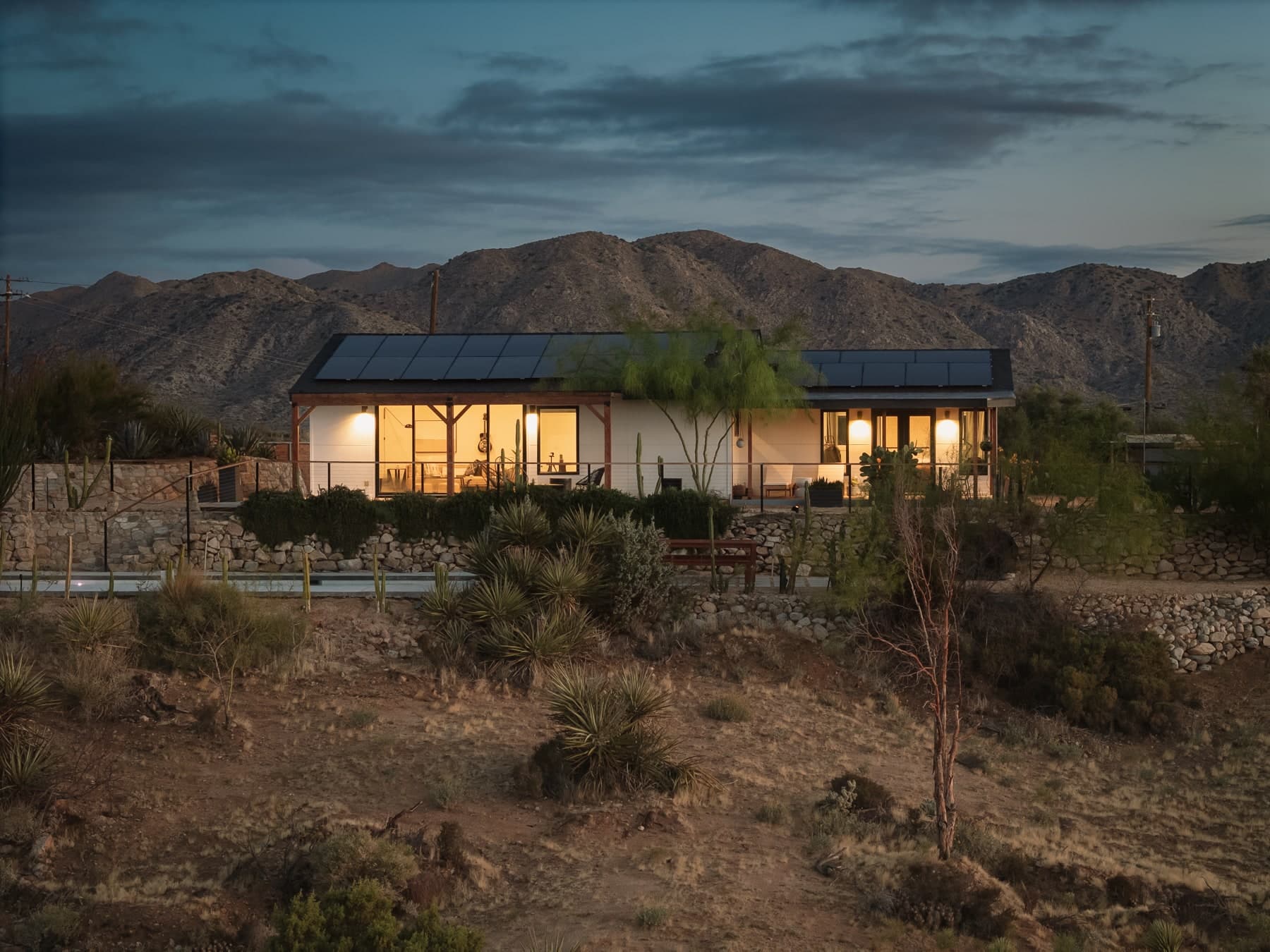 Low desert home glowing at dusk beneath mountains and a clouded sky