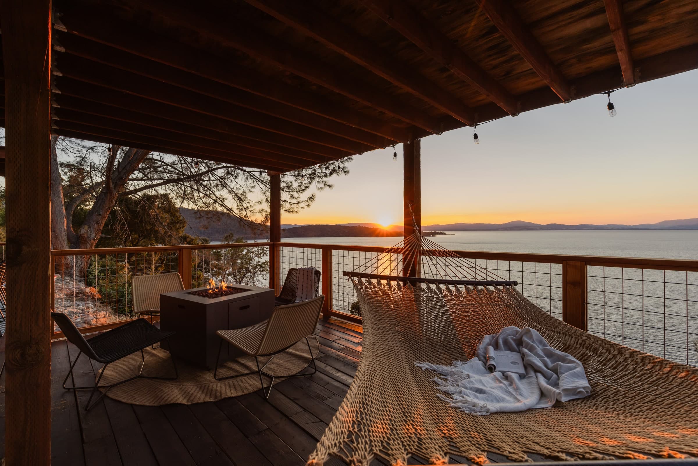 Covered lakeside deck with a rope hammock, fire pit, and lounge chairs facing the sunset