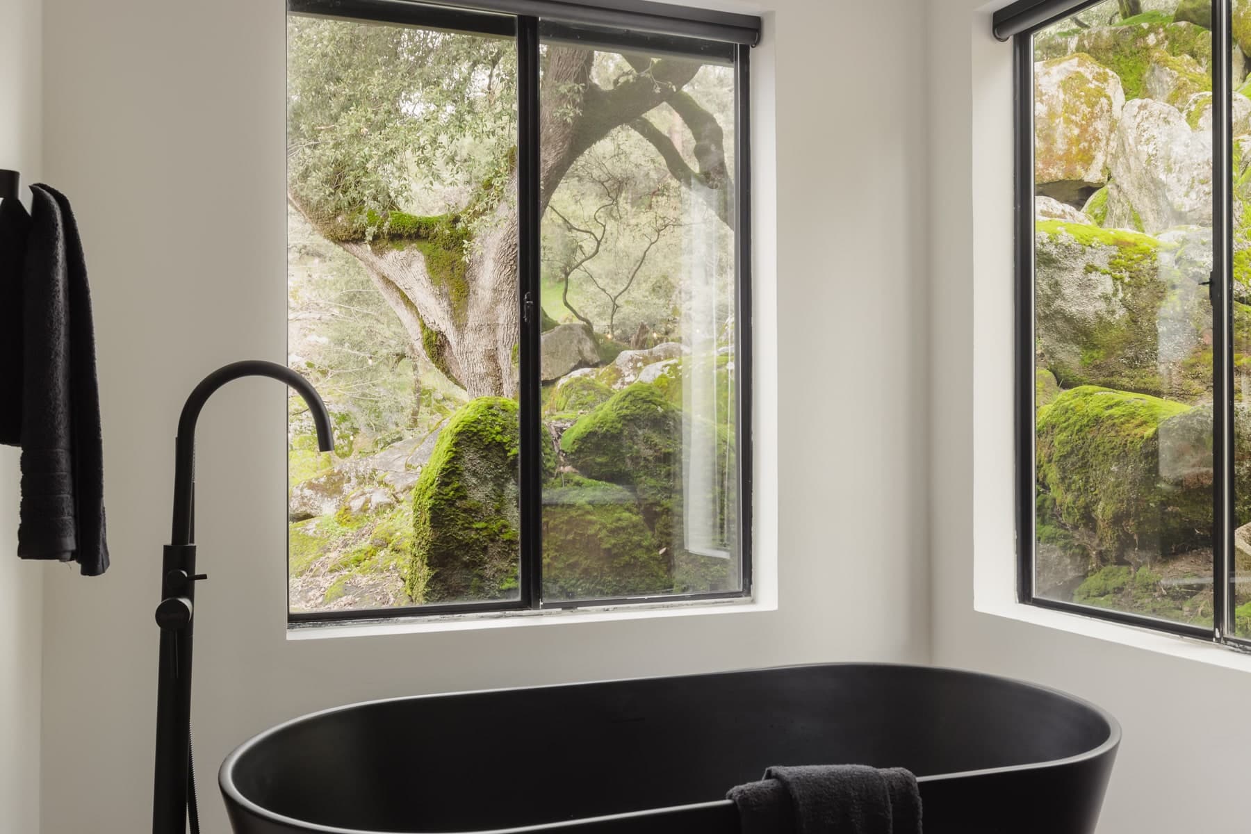 Matte black soaking tub beside large windows overlooking moss-covered boulders and oaks