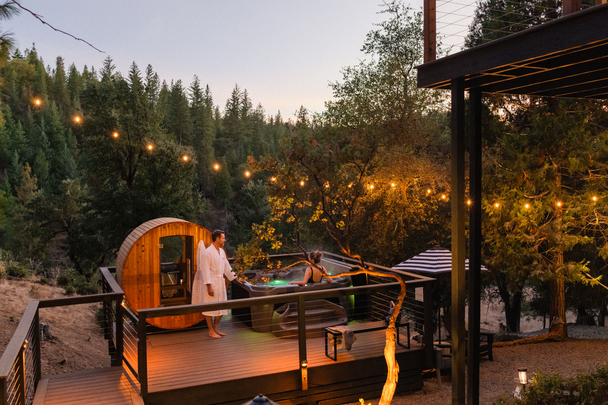 A couple at the hot tub at dusk, the barrel sauna beside them, string lights in the pines